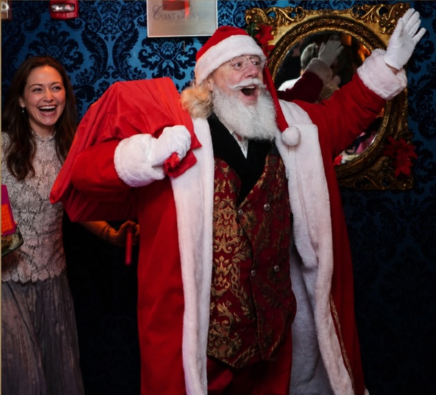 Santa James entering a celebration with a sack over his shoulder and one arm raised.